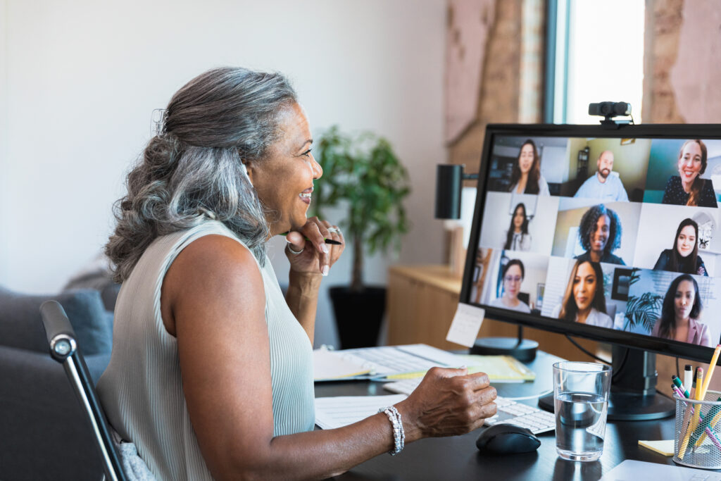The senior adult female CEO, sitting at her desk in her home office, smiles at the multi-ethnic group of employees.