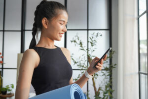 Asian woman standing holding yoga mat beside body and using smartphone to look at Castlight healthcare navigation app
