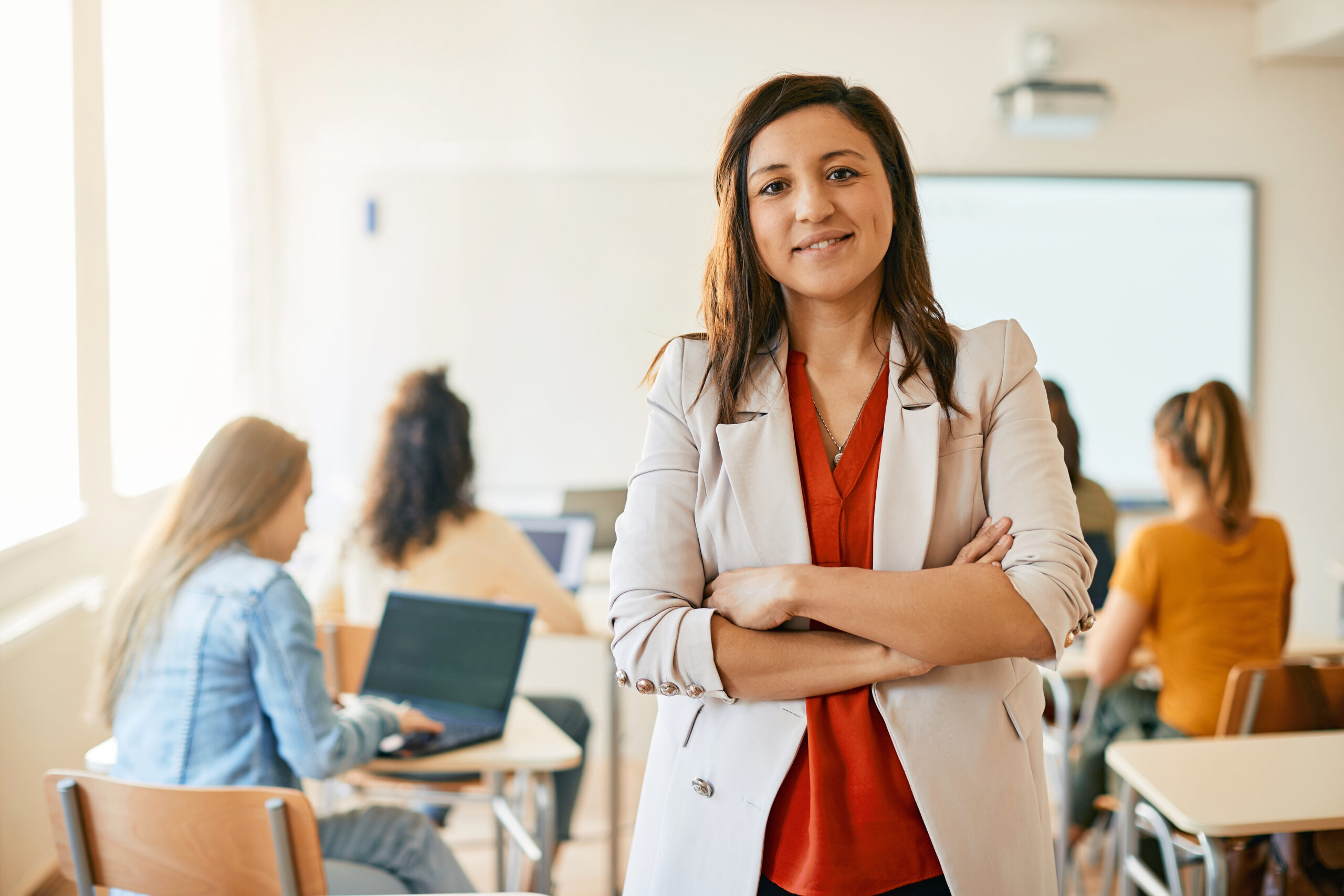 Happy female professor standing with arms crossed during computer class and looking at camera. Her students are e-learning in the background.