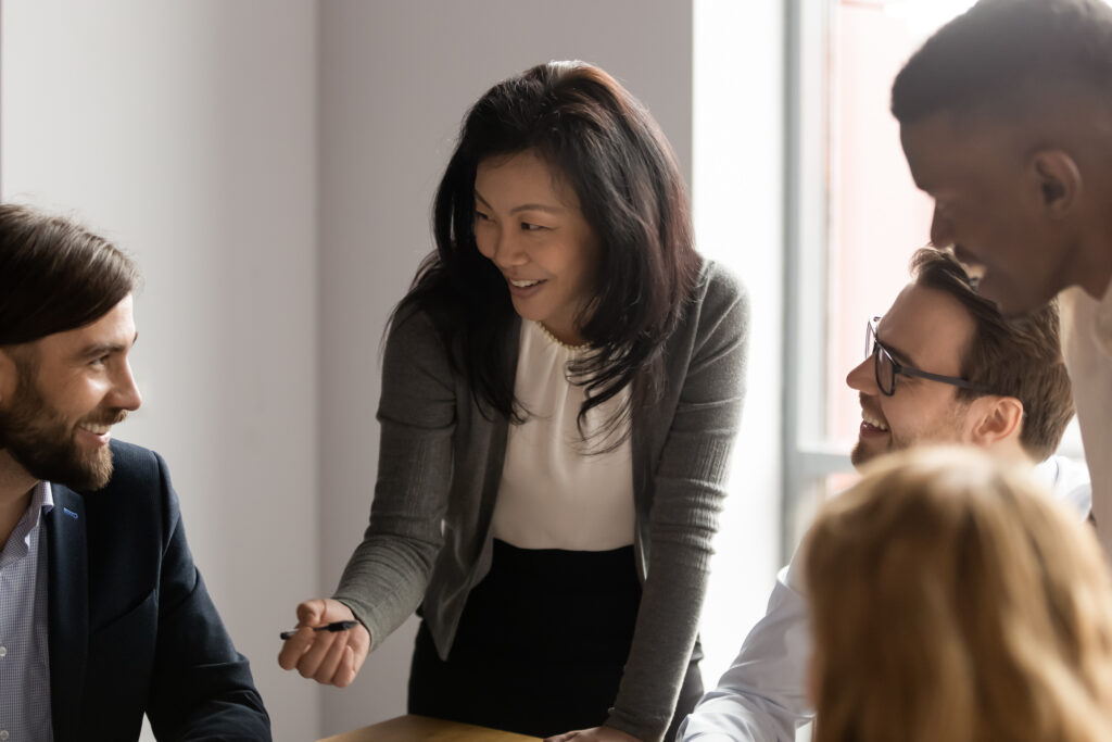 Middle aged Asian businesswoman talking to diverse colleagues at meeting, discussing project strategy and statistics, coach mentor speaking, training staff, happy business team engaged in teamwork